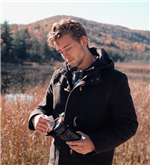 Kevin cleans a camera lens, standing in a field